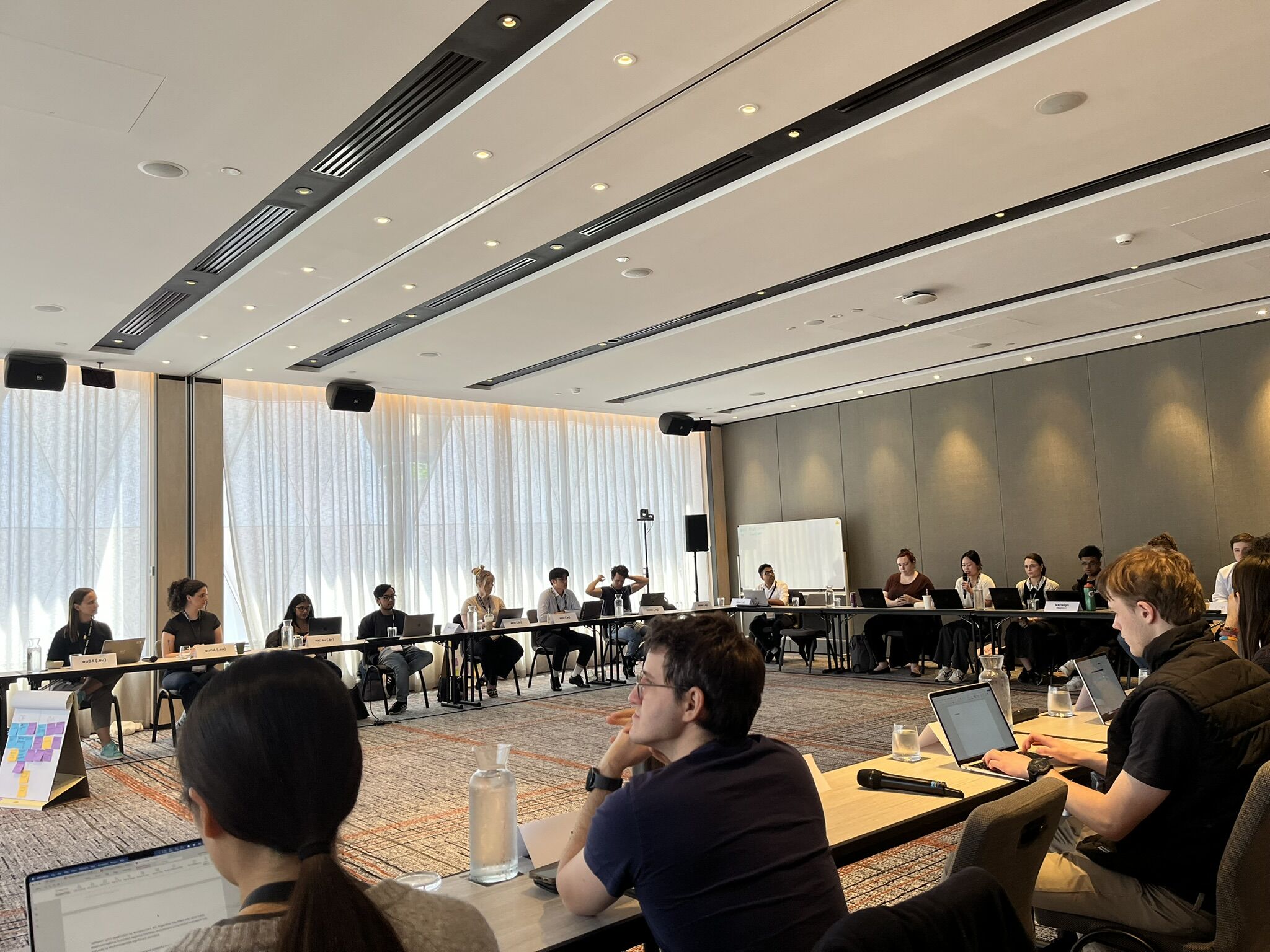 A picture of a large rectangular room with tables lining the perimeter of the room. Participants are sitting at their respective desks.