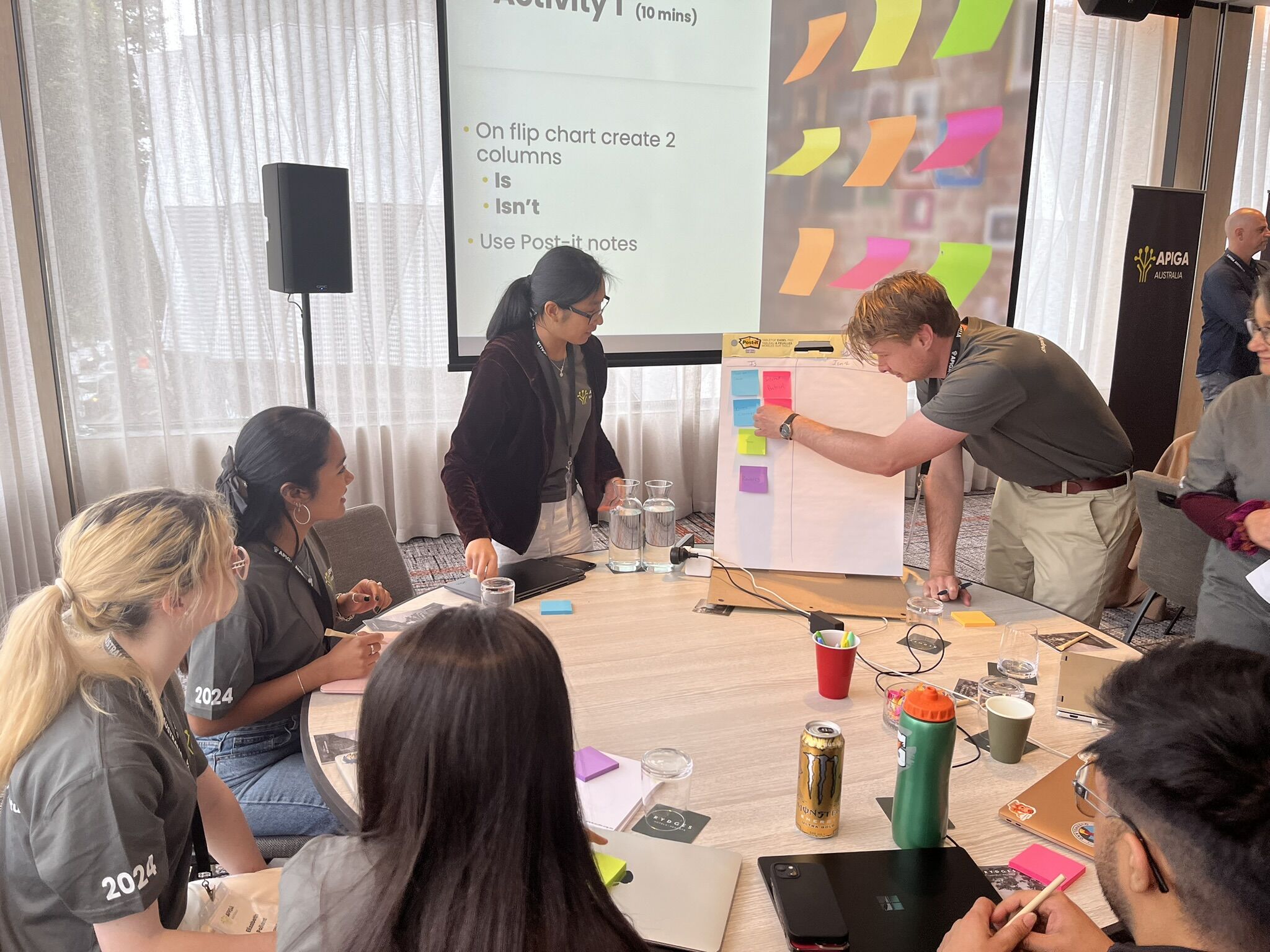 A picture of a group of participants sitting around a desk, placing sticky notes into two columns. One column is labelled 'is' and the other is labelled 'isn't'. The activity was run during Adam's talk and participants had to classify whether items were part of the internet or not.
